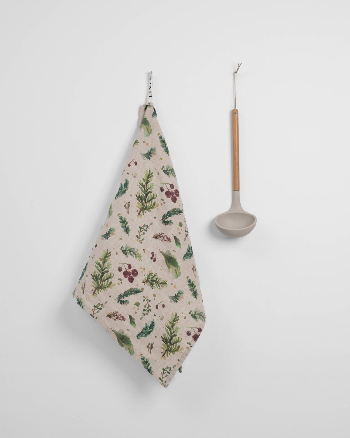 A beige kitchen tea towel with a green and burgundy botanical pattern hangs on a white wall next to a matte beige ladle with a light wood handle.