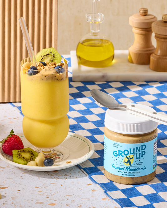 Yellow smoothie with fruit and a jar of Ground Up toasted macadamia nut butter on a checkered tablecloth.