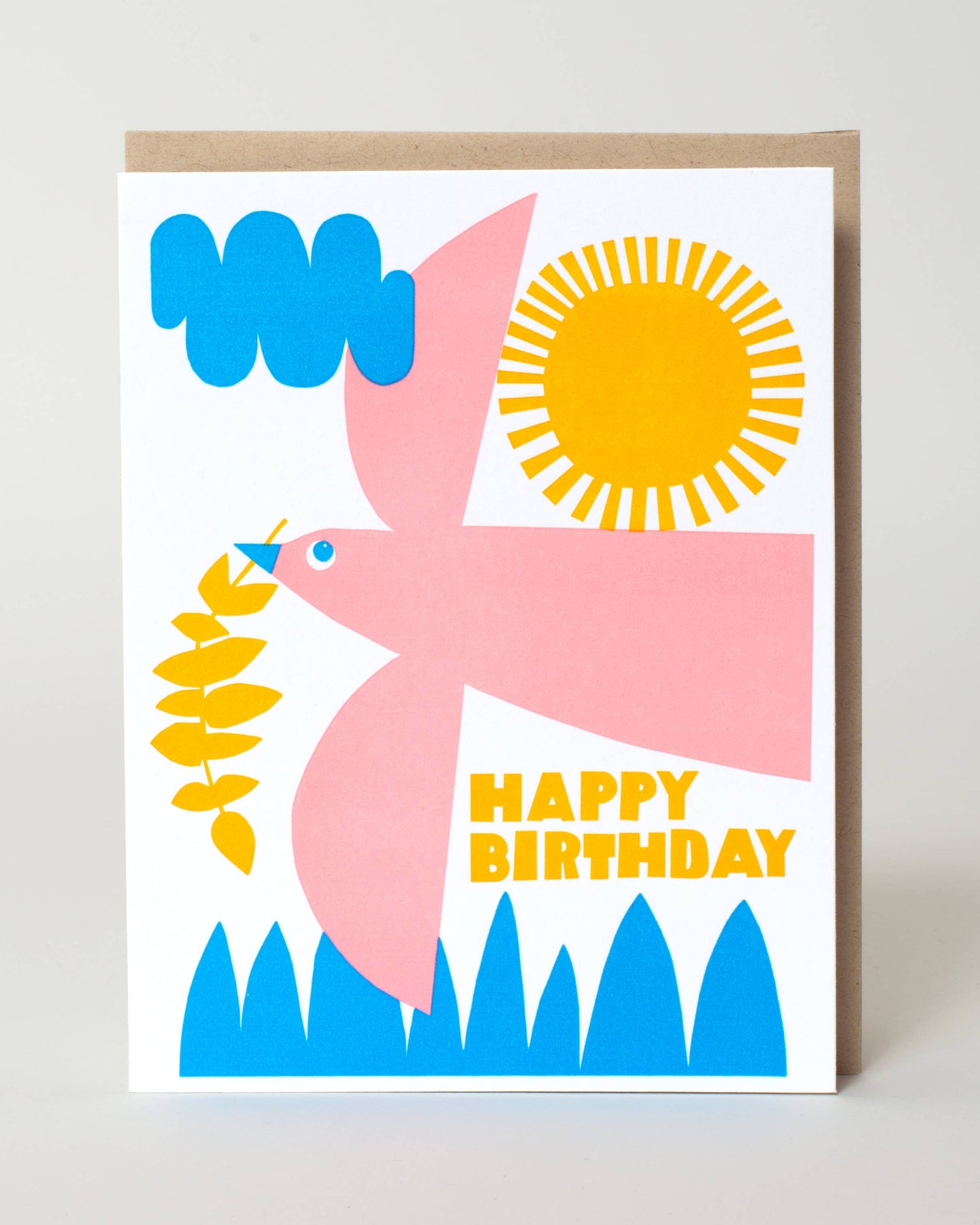 A square birthday greeting card with a minimalist flat illustration of a pink bird, blue clouds and grass, a yellow sun, and the words “HAPPY BIRTHDAY” in yellow, shown standing against a light neutral background.