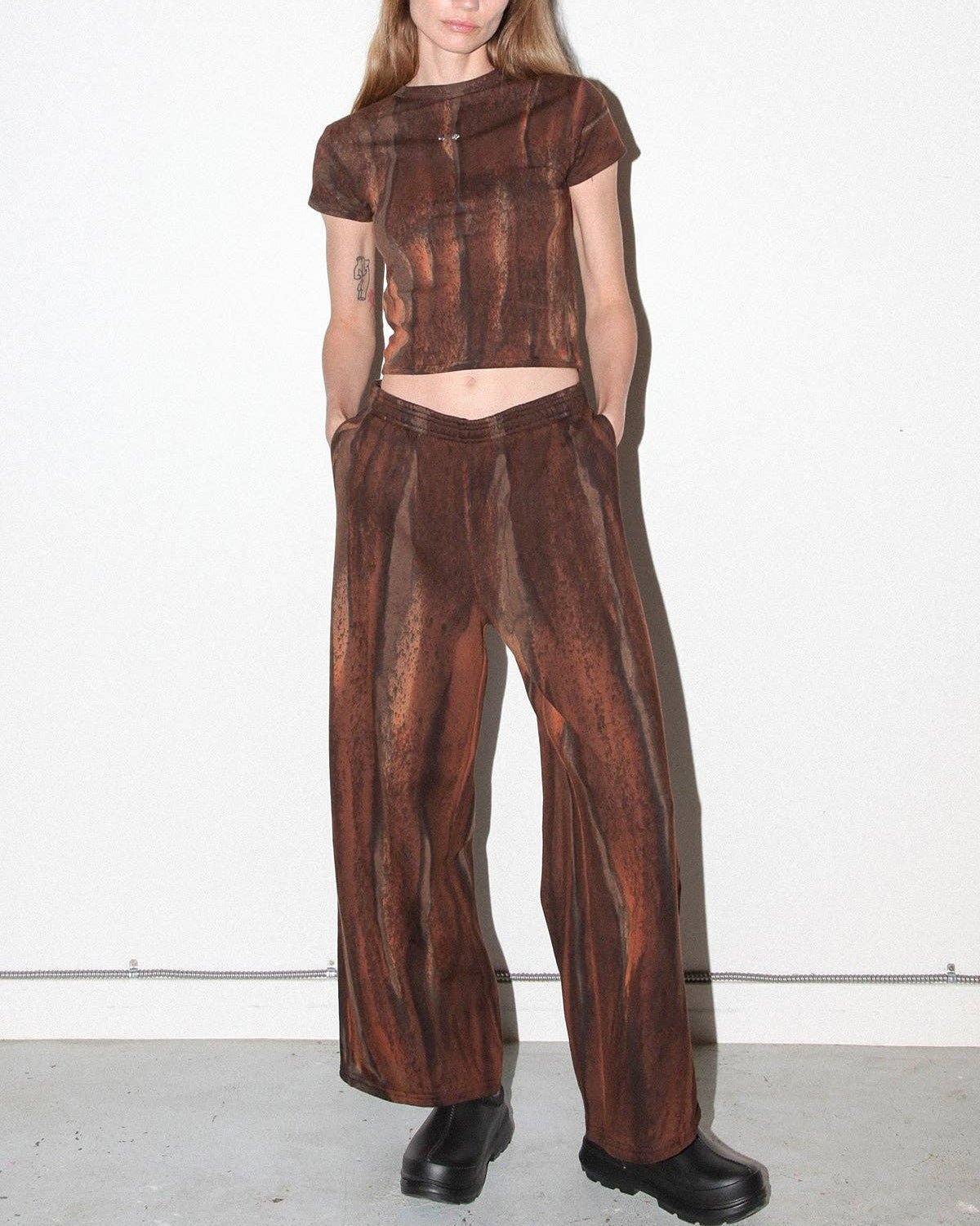 A model stands against a plain white wall wearing a matching brown textured two-piece outfit consisting of a short-sleeve cropped top and wide-leg pants, paired with black boots.