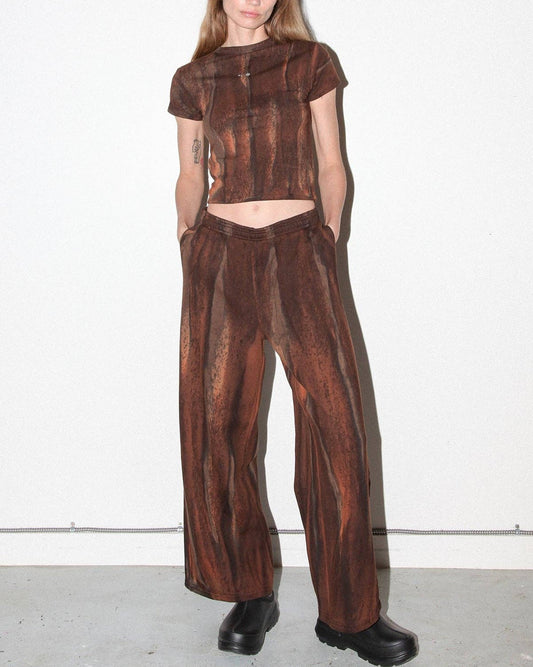 A model stands against a plain white wall wearing a matching brown textured two-piece outfit consisting of a short-sleeve cropped top and wide-leg pants, paired with black boots.
