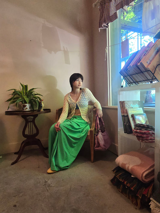 Person wearing a green dress and patterned cardigan in a room with a window and furniture.