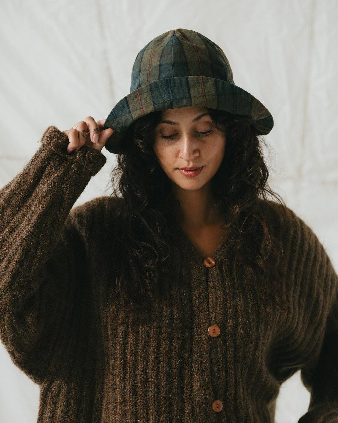 Woman wearing a plaid hat and brown knitted cardigan against a white background