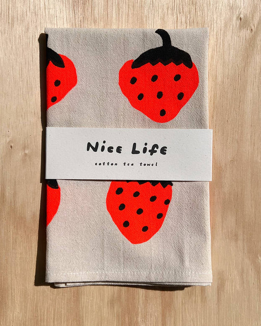 Tea towel with strawberry design and 'Nice Life' label on a wooden surface