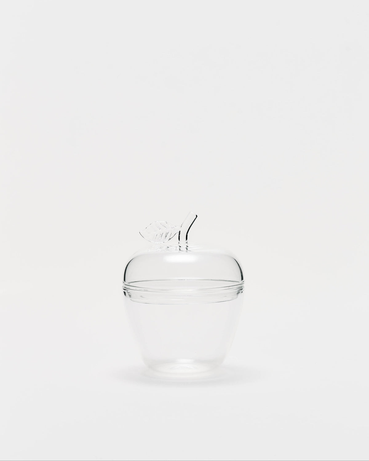 A clear glass lidded container shaped like an apple, featuring a small glass stem handle on top, set against a solid light gray background.