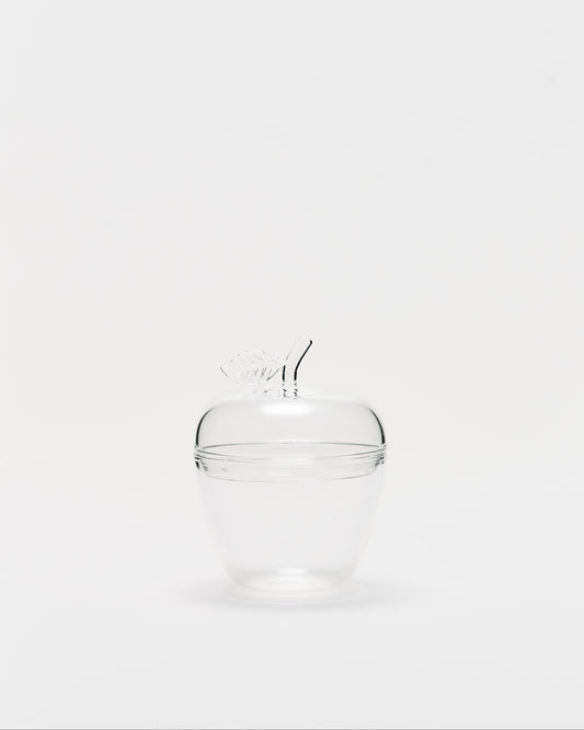 A clear glass lidded container shaped like an apple, featuring a small glass stem handle on top, set against a solid light gray background.