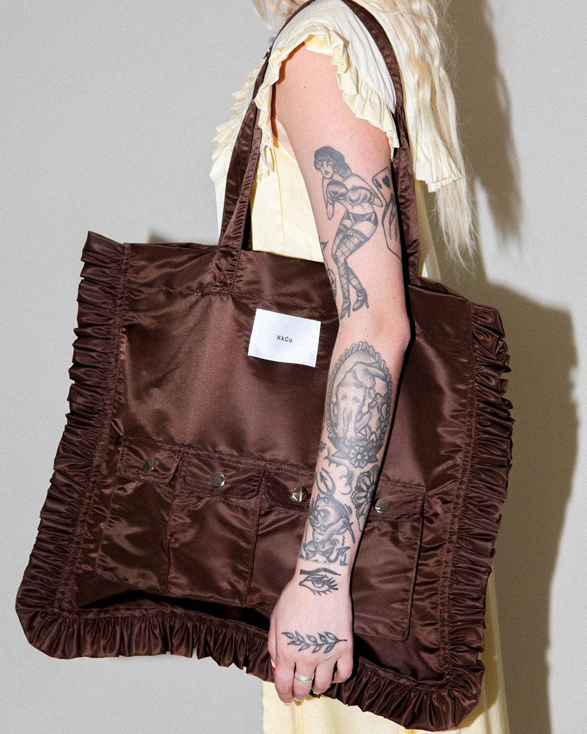 A blonde model wearing a pale yellow dress carries a large brown satin tote bag with ruffled trim and multiple front pockets. The model has visible tattoos on her arm, and the background is a plain light gray wall.