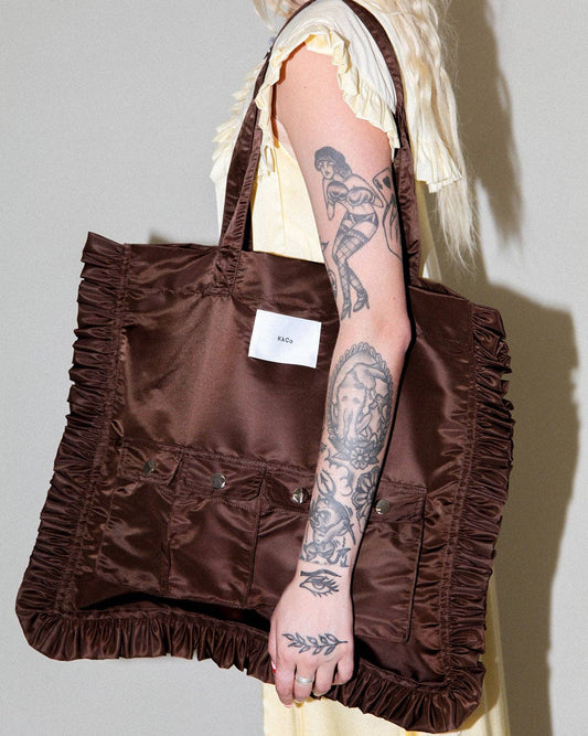 A blonde model wearing a pale yellow dress carries a large brown satin tote bag with ruffled trim and multiple front pockets. The model has visible tattoos on her arm, and the background is a plain light gray wall.
