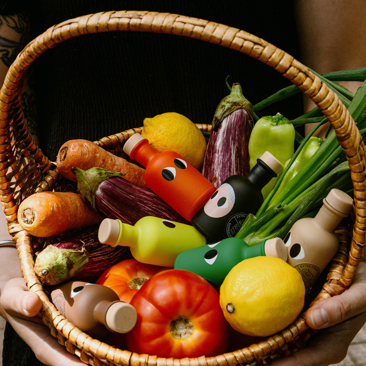 A woven wicker basket filled with colorful cartoon-style vegetable and bottle toys, including orange, green, red, black, and beige pieces with simple facial features, held by hands against a dark background.