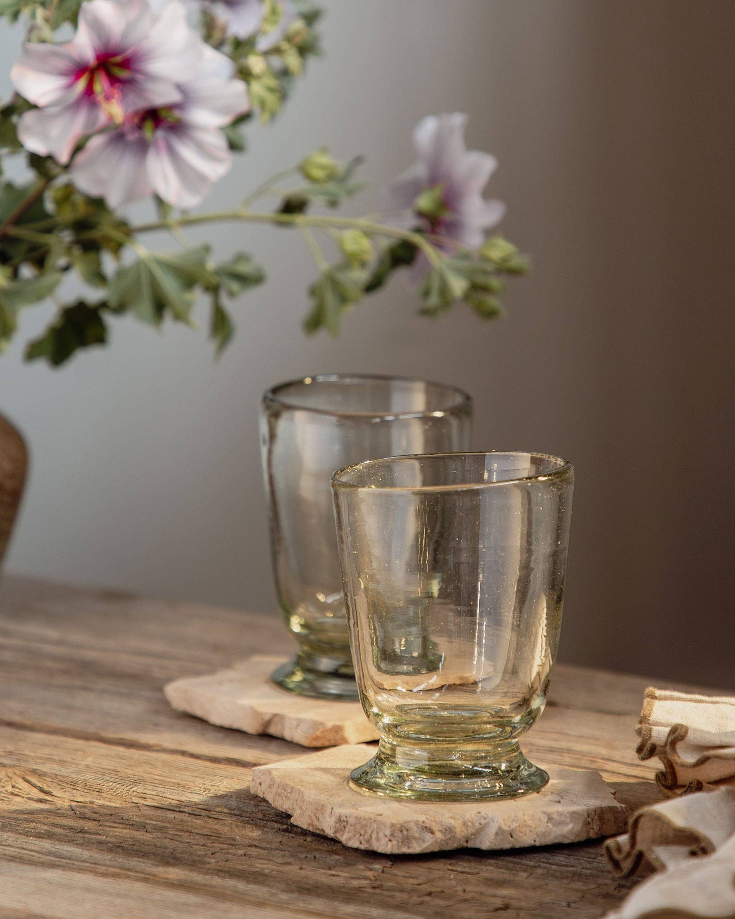 Two glass tumblers on a wooden coaster with flowers in the background