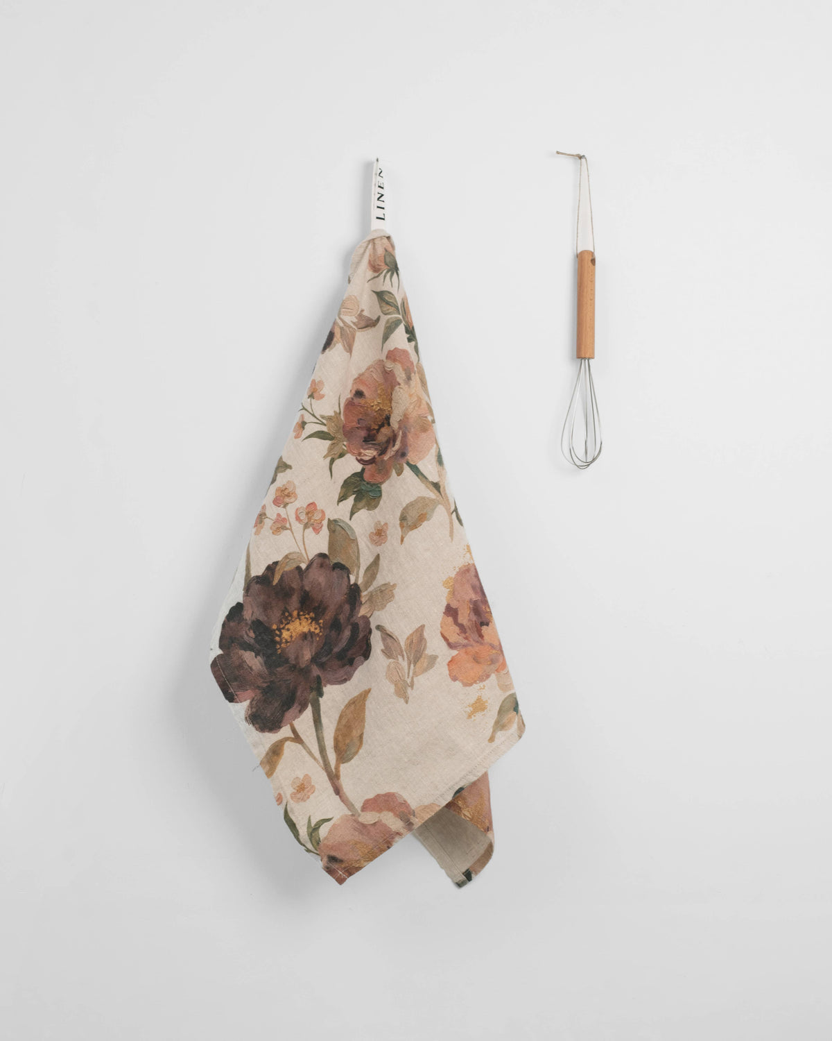 A floral kitchen towel with a cream background and dark brown, orange, and green botanical print hangs from a metal hook next to a small whisk with a light wood handle.