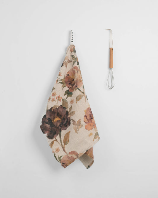A floral kitchen towel with a cream background and dark brown, orange, and green botanical print hangs from a metal hook next to a small whisk with a light wood handle.