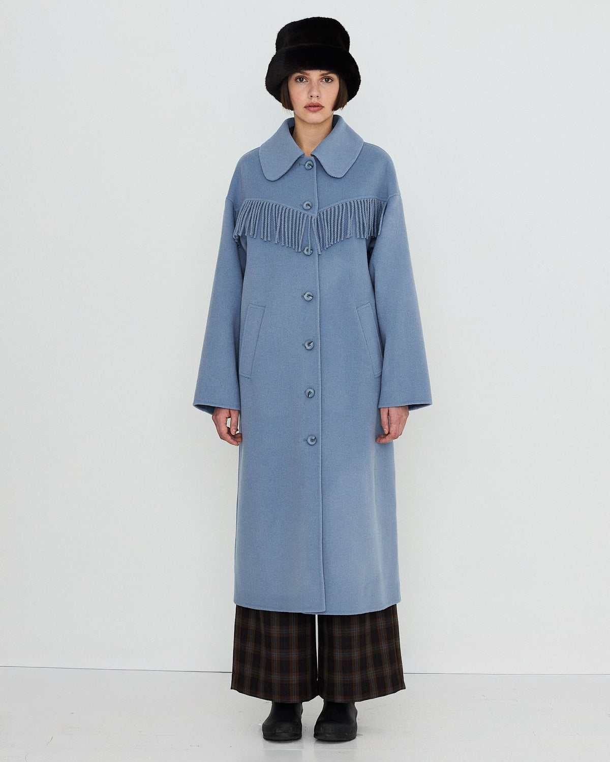 A model stands against a plain white studio background wearing a long light blue coat with a wide collar and dark fringe detail across the chest, paired with dark plaid pants, black shoes, and a black hat.