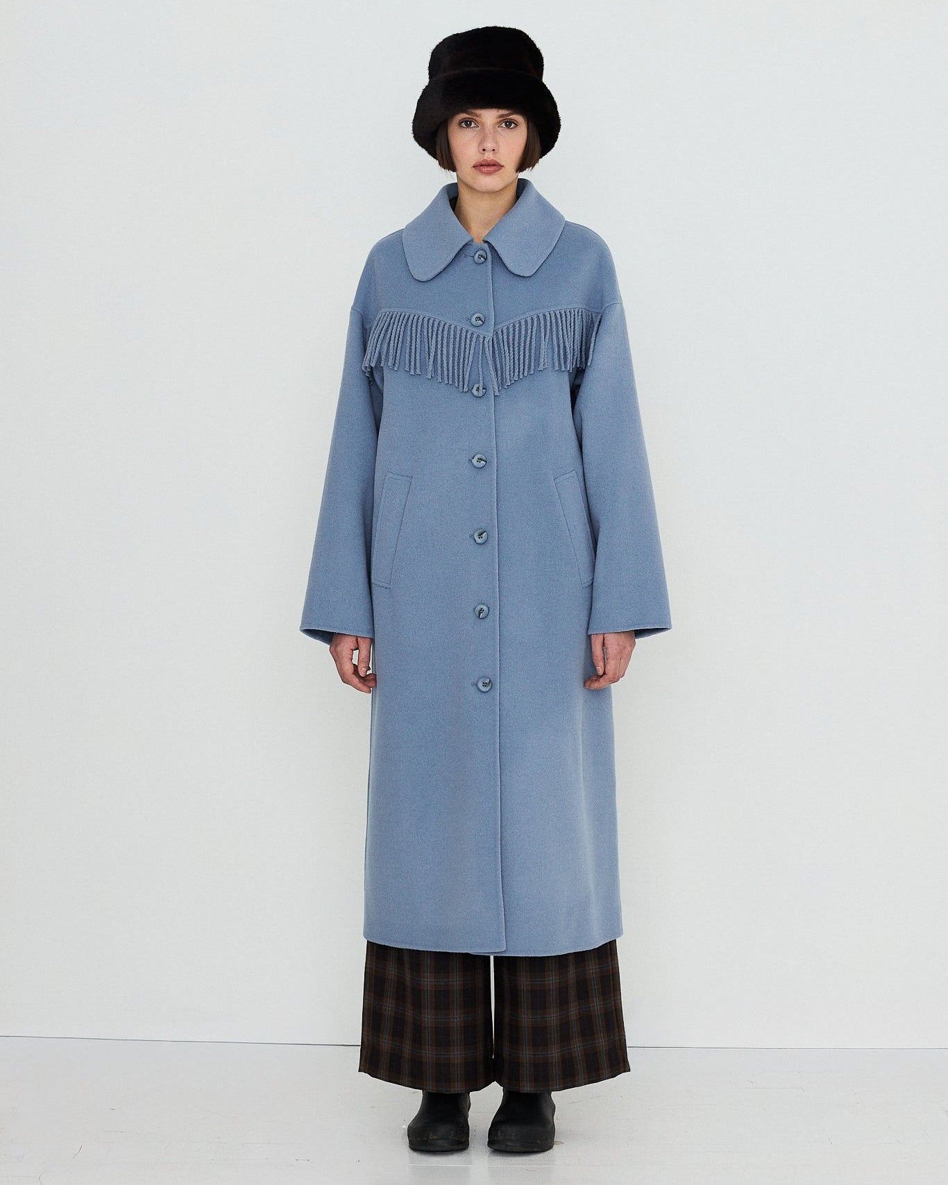 A model stands against a plain white studio background wearing a long light blue coat with a wide collar and dark fringe detail across the chest, paired with dark plaid pants, black shoes, and a black hat.