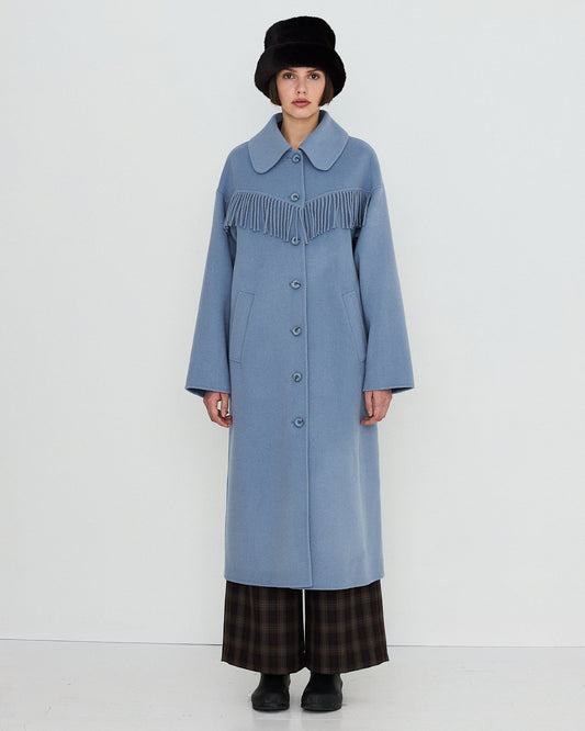 A model stands against a plain white studio background wearing a long light blue coat with a wide collar and dark fringe detail across the chest, paired with dark plaid pants, black shoes, and a black hat.