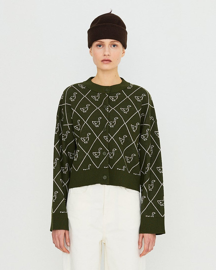 A model stands against a plain white studio background wearing a dark green long-sleeve knit sweater with a white geometric pattern, white trousers, a dark beanie, and dark brown shoes.