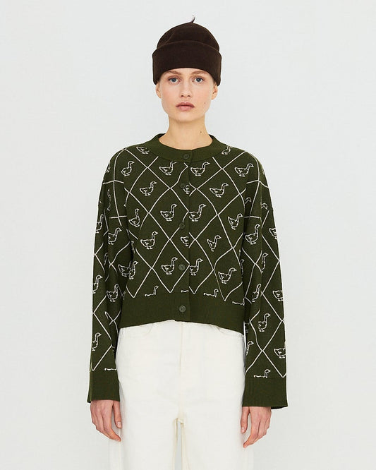 A model stands against a plain white studio background wearing a dark green long-sleeve knit sweater with a white geometric pattern, white trousers, a dark beanie, and dark brown shoes.