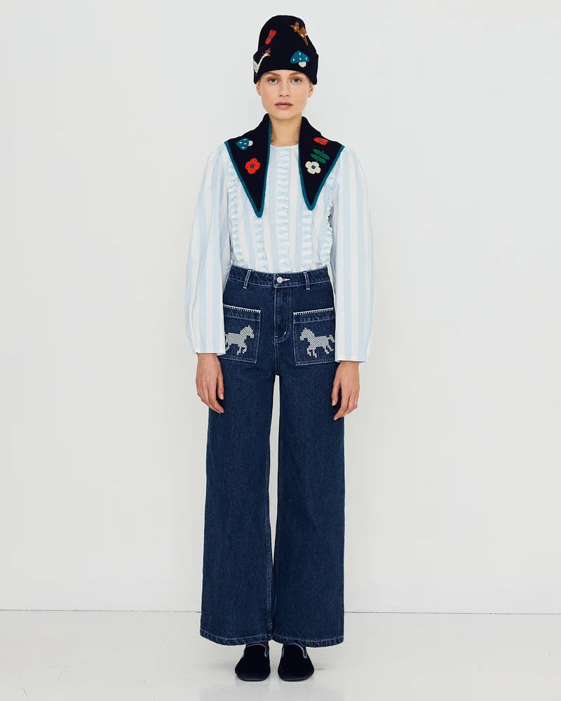 A model stands against a white studio background wearing a light blue long-sleeve top with dark green embroidered yoke details, dark blue wide-leg denim pants with embroidered horse motifs on the pockets, and a dark knit beanie with colorful embroidery.