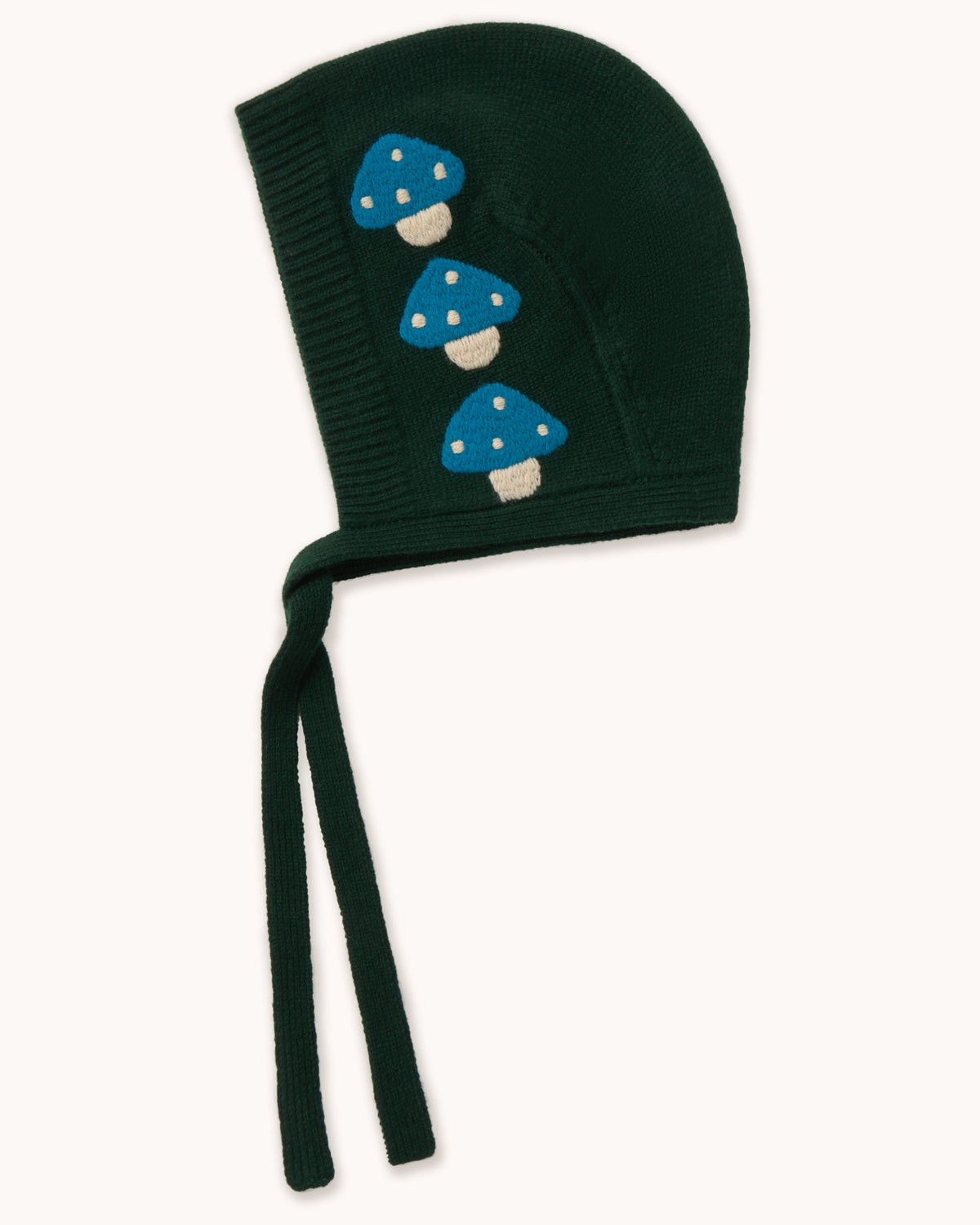 A dark forest green knit bonnet with three embroidered blue and white mushrooms on the side, featuring long ties, shown against a plain off-white background.