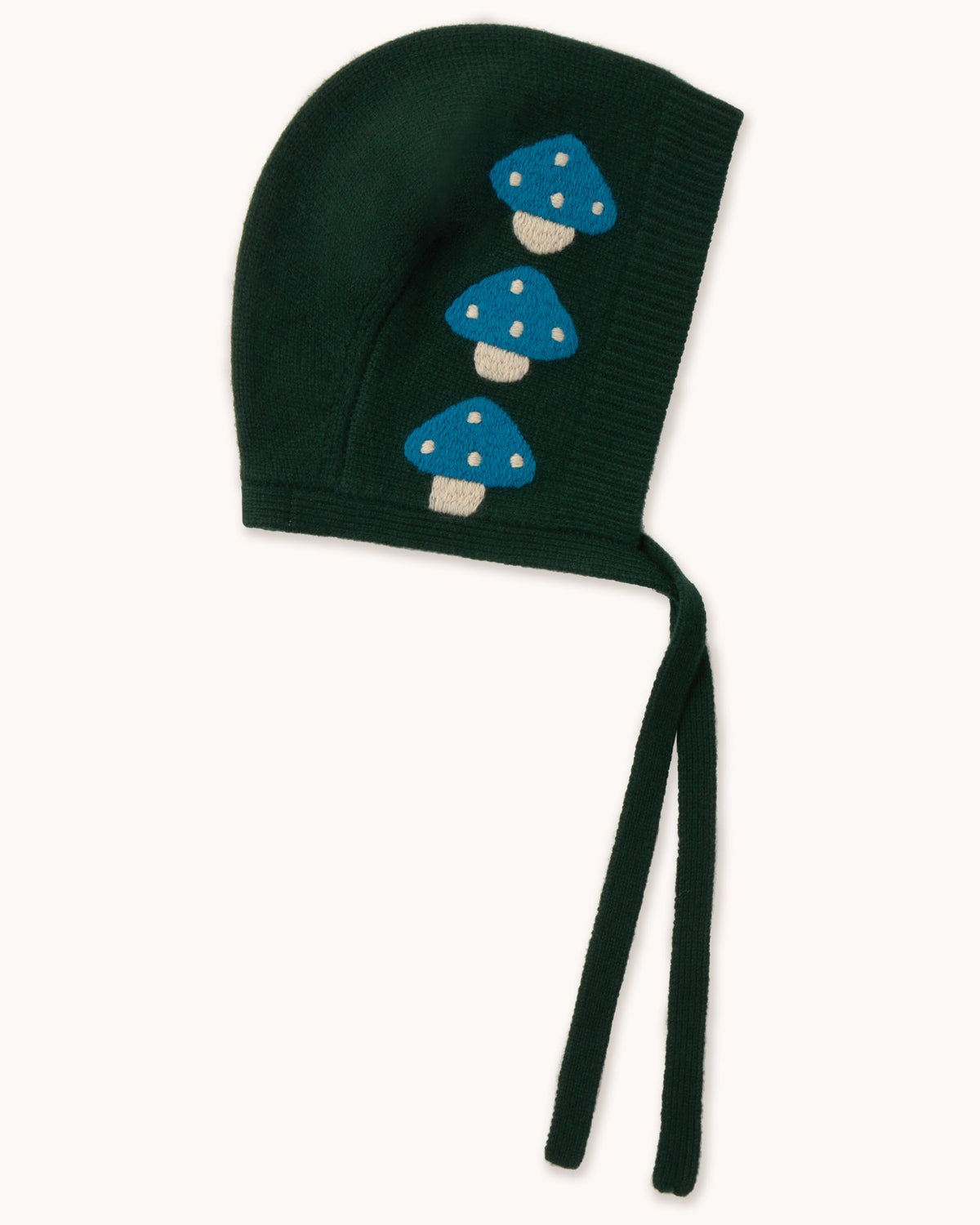 A dark forest green knit bonnet with three embroidered blue and white mushrooms on the side, featuring long ties, shown against a plain off-white background.
