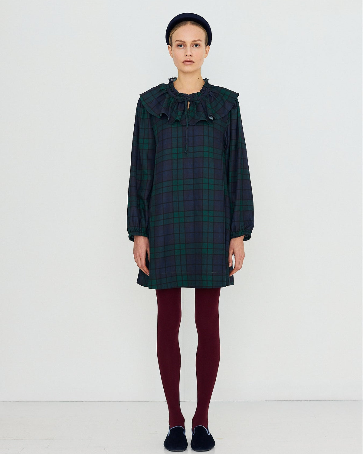 A model stands against a plain white studio background wearing a dark green and black plaid long-sleeve dress with a large ruffled collar, paired with burgundy tights and dark slip-on shoes.