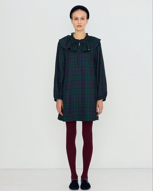 A model stands against a plain white studio background wearing a dark green and black plaid long-sleeve dress with a large ruffled collar, paired with burgundy tights and dark slip-on shoes.