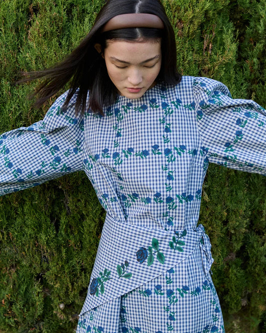 A woman wearing a blue and white gingham dress with green floral embroidery, posing outdoors against a dense green hedge.