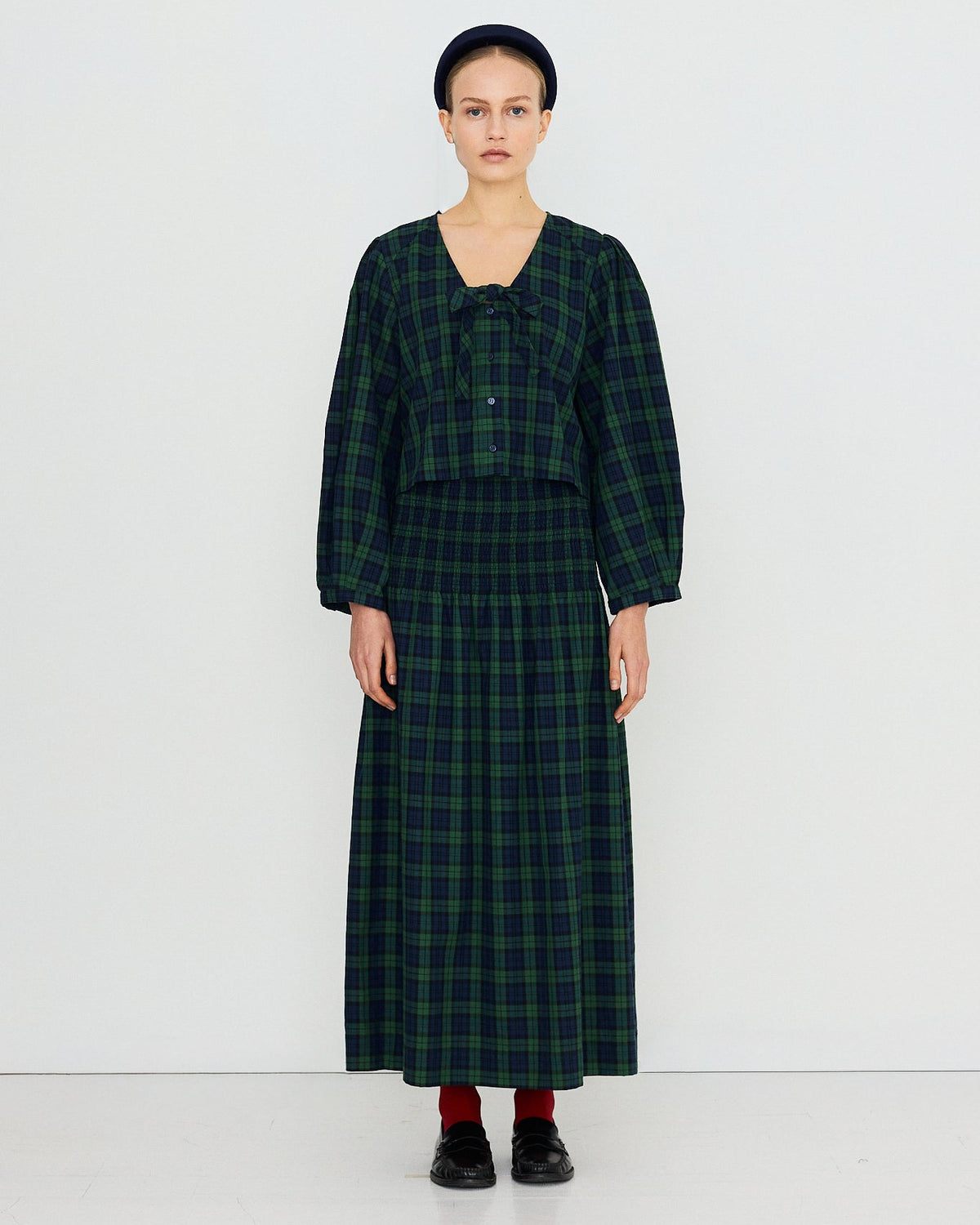 A model stands against a plain white studio background wearing a dark green and black plaid two-piece outfit consisting of a cropped long-sleeve top with a bow detail and a matching long skirt.
