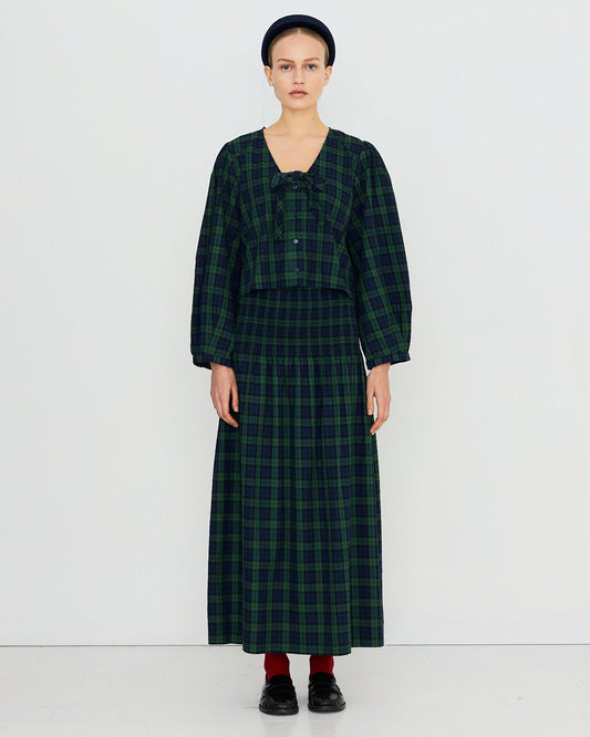 A model stands against a plain white studio background wearing a dark green and black plaid two-piece outfit consisting of a cropped long-sleeve top with a bow detail and a matching long skirt.