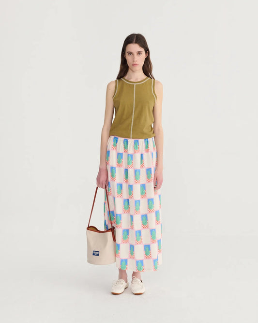 Woman wearing a green sleeveless top and patterned skirt holding a beige bag on a white background