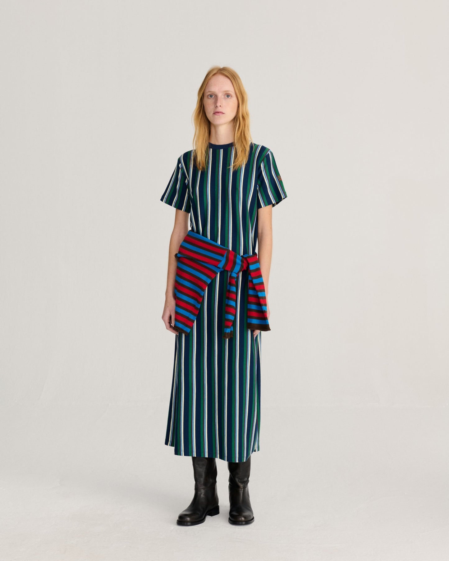 A woman stands against a light gray studio background wearing a short-sleeve midi dress with vertical dark green, teal, and white stripes, a red and navy striped wrap tied at the waist, and black ankle boots.