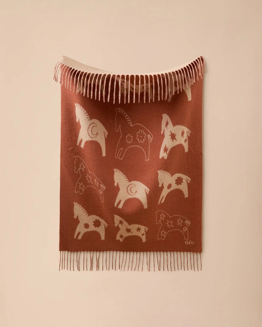 A rust-brown woven throw blanket featuring cream-colored Swedish Pallas deer motifs and fringed edges, displayed against a solid beige wall.