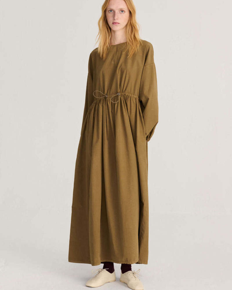 A woman modeling a long-sleeved, ankle-length olive green dress with a drawstring waist, standing against a plain light gray studio background.