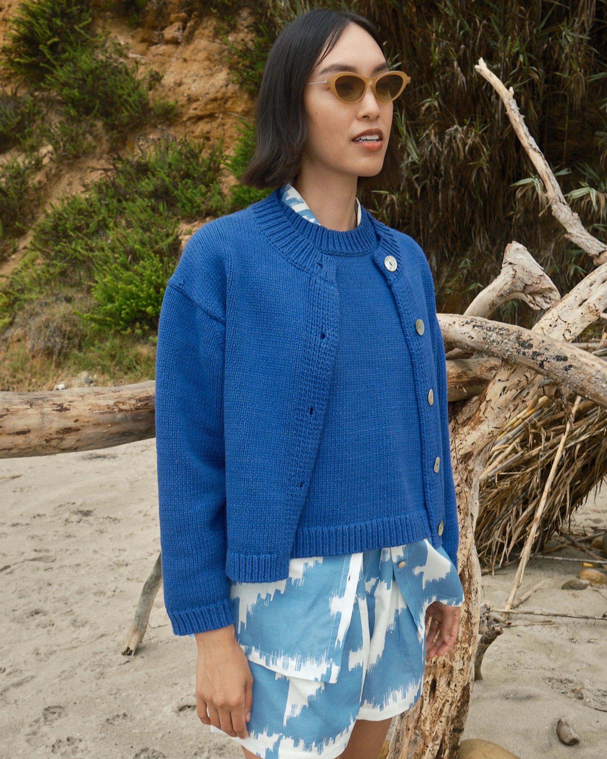 Woman in a blue cardigan and patterned shorts standing on a beach with driftwood and greenery.