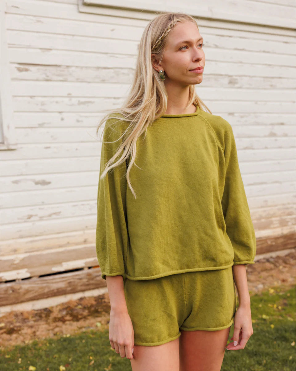 A woman wearing a matching olive green lounge set consisting of a long-sleeve top and shorts, standing outdoors in front of a white wooden wall.