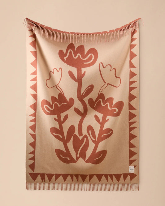 A beige woven throw blanket with a terracotta floral pattern and fringed edges, hanging against a solid beige wall.