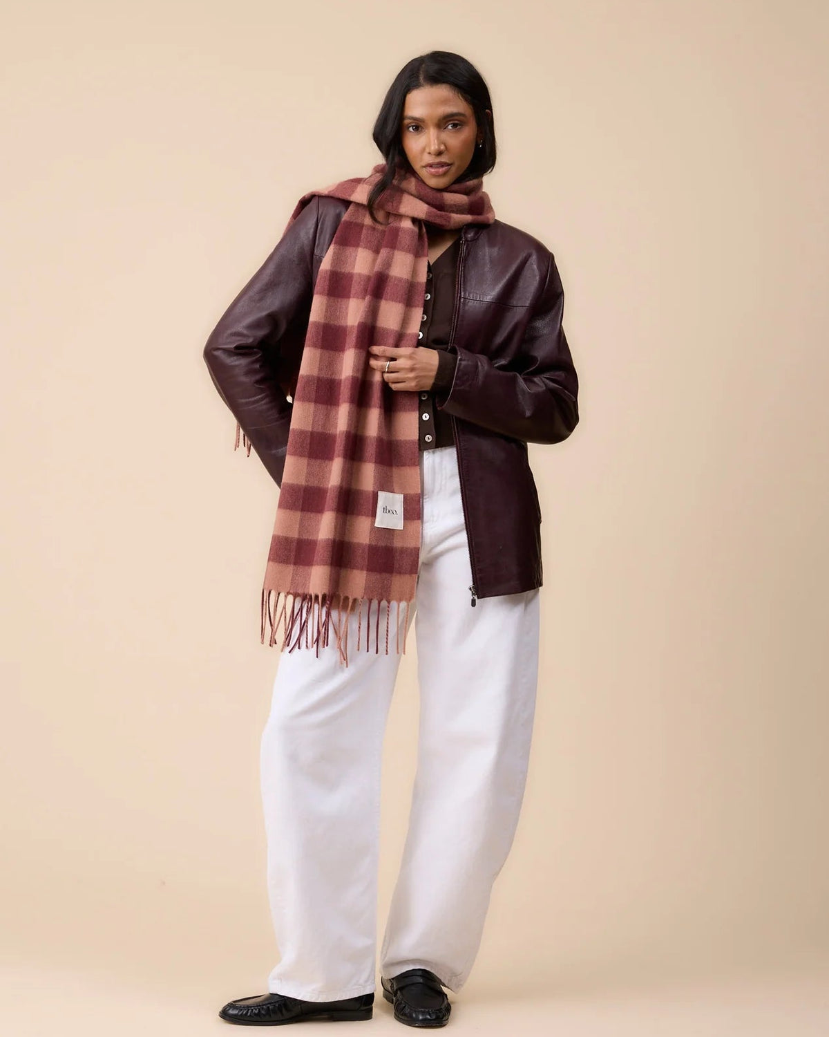 A model stands against a solid beige studio background wearing a dark brown leather jacket, white wide-leg trousers, black shoes, and a rust-brown and beige plaid scarf.