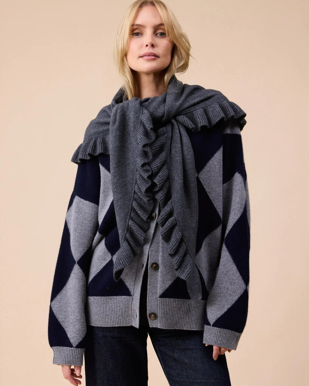 A woman modeling a dark gray knit cardigan with a geometric navy and gray diamond pattern, styled with a matching dark gray ruffled scarf, against a solid beige background.