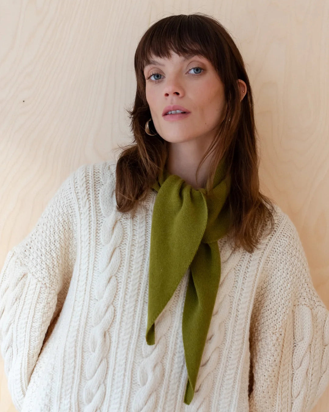 A woman with brown hair and bangs wearing a cream cable-knit sweater and an olive green bow tie, standing against a light wood-paneled wall.