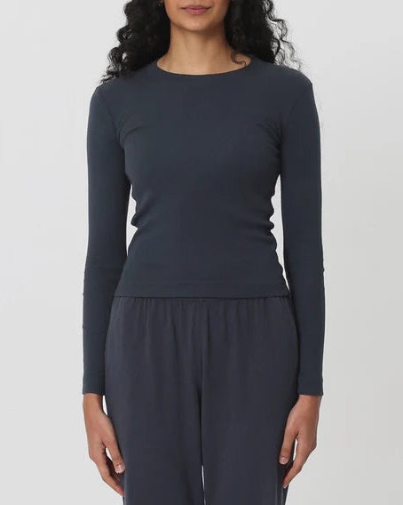 A woman with long dark curly hair stands against a light gray studio background wearing a dark slate blue long-sleeve crewneck top and matching dark pants.