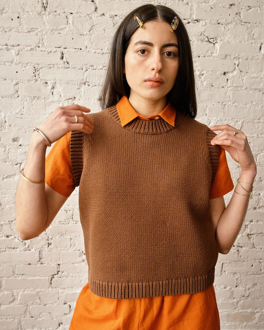 Woman wearing a brown knitted vest over an orange shirt and skirt against a white brick wall.