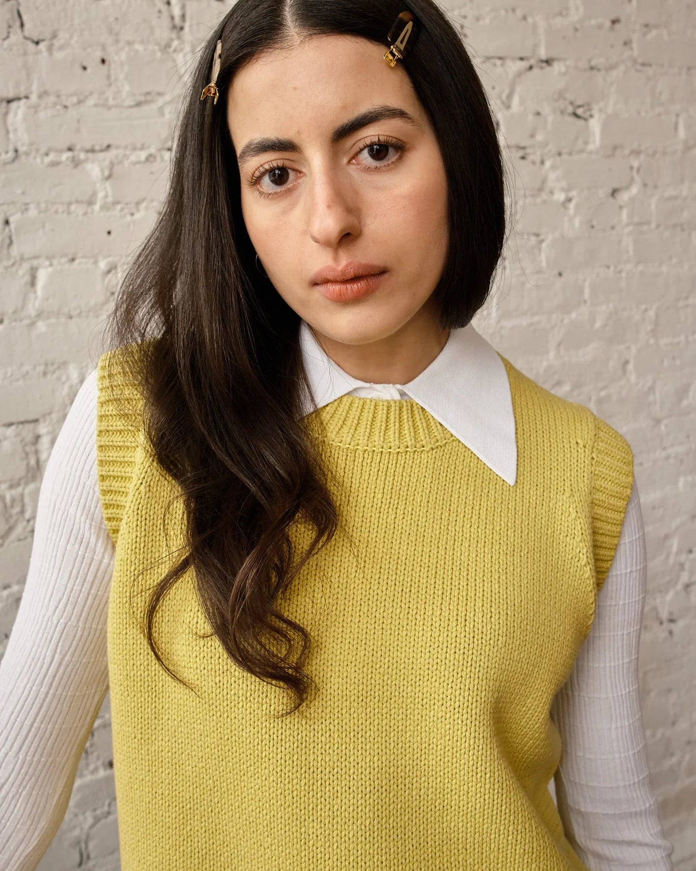 Woman wearing a yellow knitted vest over a white shirt against a brick wall.