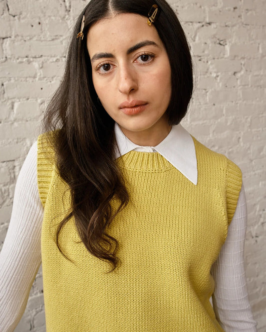 Woman wearing a yellow knitted vest over a white shirt against a brick wall.