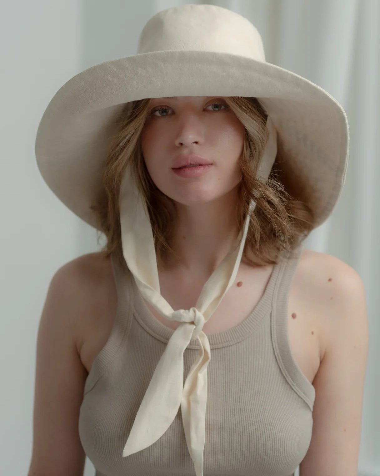 Woman wearing a beige sun hat with a scarf, standing against a plain background