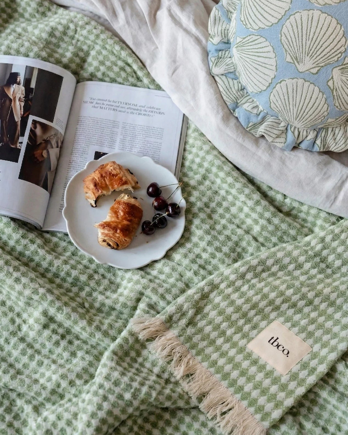 Green textured blanket with a plate of food, magazine, and decorative pillow on a bed.