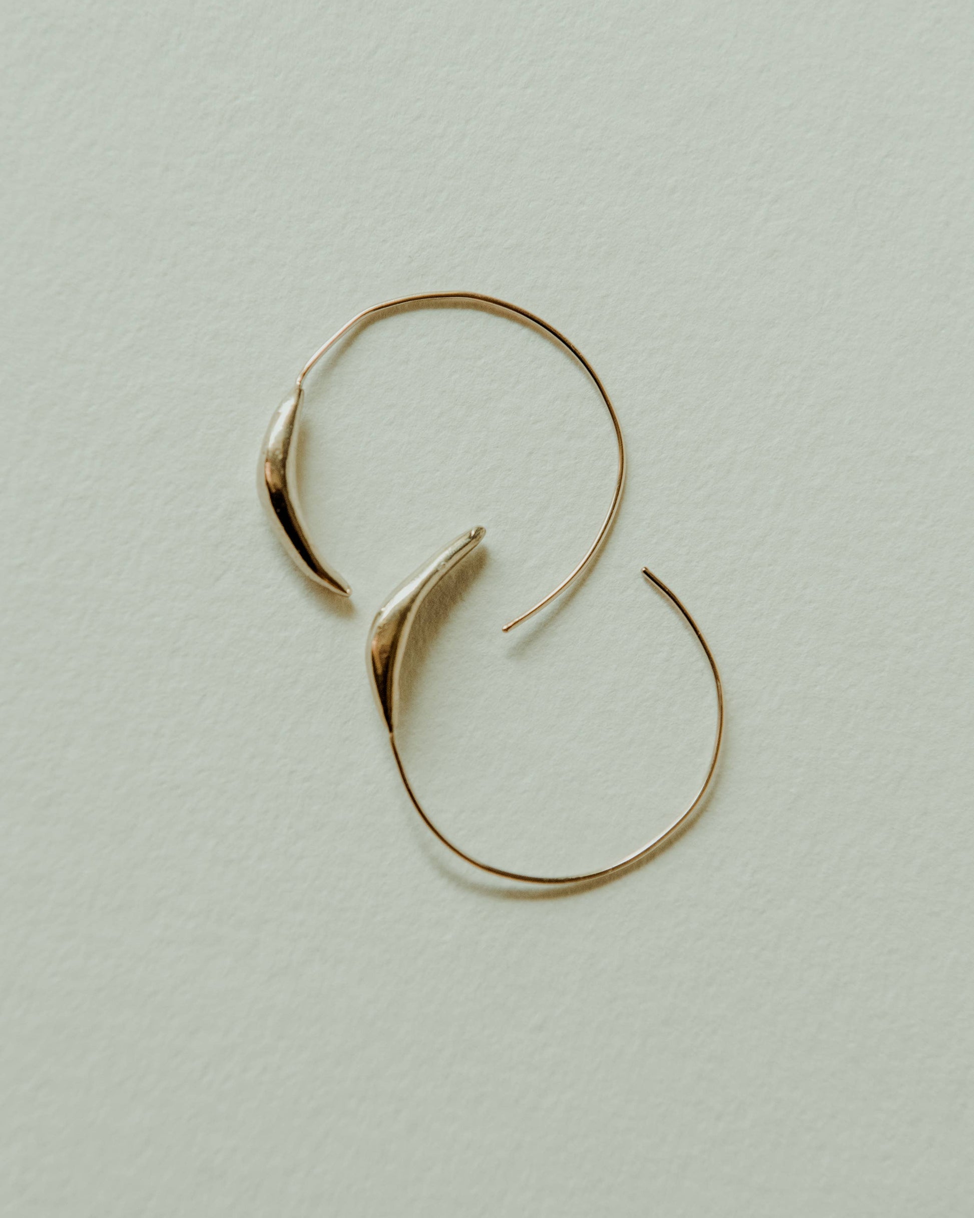 A pair of thin gold hoop earrings with organic, leaf-like curved tips, displayed on a textured off-white background.