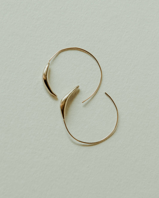A pair of thin gold hoop earrings with organic, leaf-like curved tips, displayed on a textured off-white background.
