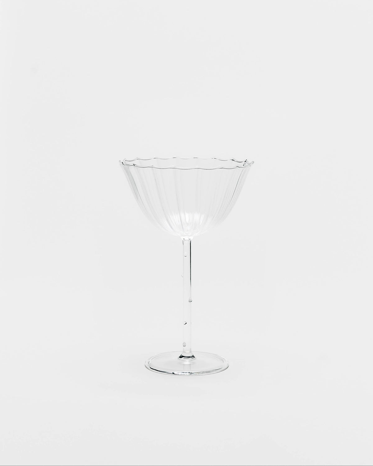 A clear glass cocktail coupe with a fluted rim and a slender stem, photographed against a seamless light gray background.