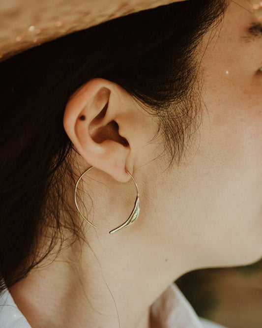A close-up side profile of a person wearing a delicate gold hoop earring with a curved leaf or feather detail hanging from the lower rim.