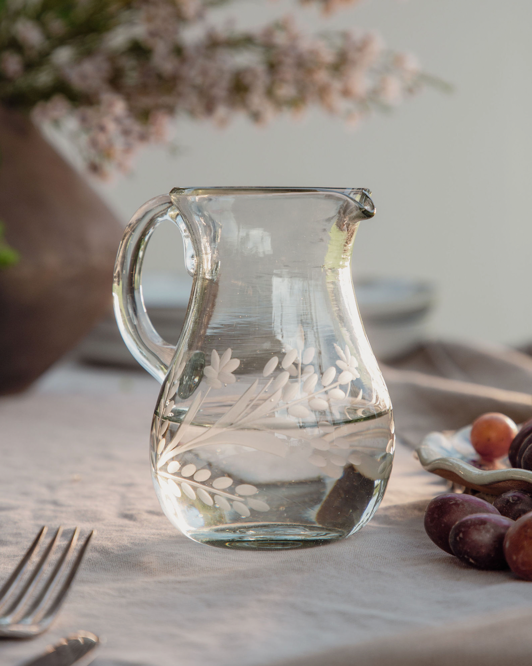 Clear glass pitcher with engraved design on a table with flowers and fruits in the background